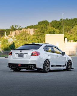 A white sedan with aftermarket modifications is parked on a smooth gray surface. The car features a sporty design with a custom rear diffuser, dual exhausts, tinted windows, and distinctive black wheel rims. Green trees and a cityscape with buildings are visible in the background under a clear blue sky.