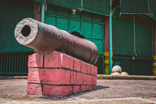 An old cannon is mounted on a pink brick base in an outdoor setting. The background features a dark green building facade with vertical wooden blinds and pillars. The ground is paved, and three cannonballs are lying nearby.