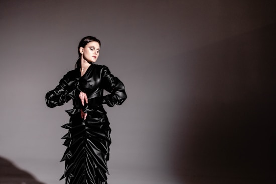 A woman is wearing an elaborate black gown with geometric, angular designs and ruffles, standing against a plain background. Her expression is contemplative, and her makeup is bold with smoky eyeshadow. The lighting creates a dramatic contrast between the subject and the background.