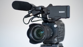 A professional video camera equipped with a large lens and an attached microphone. The camera is mounted on a tripod, and the brand name on the camera is partially visible. The focus is on the lens, which features detailed inscriptions, demonstrating the advanced features of the equipment.