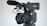A professional video camera equipped with a large lens and an attached microphone. The camera is mounted on a tripod, and the brand name on the camera is partially visible. The focus is on the lens, which features detailed inscriptions, demonstrating the advanced features of the equipment.