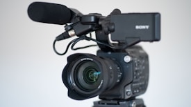 A professional video camera equipped with a large lens and an attached microphone. The camera is mounted on a tripod, and the brand name on the camera is partially visible. The focus is on the lens, which features detailed inscriptions, demonstrating the advanced features of the equipment.
