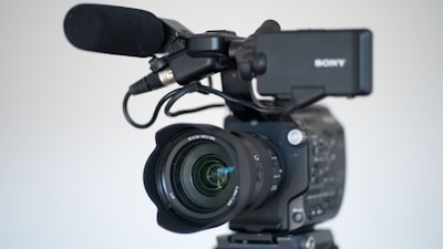A professional video camera equipped with a large lens and an attached microphone. The camera is mounted on a tripod, and the brand name on the camera is partially visible. The focus is on the lens, which features detailed inscriptions, demonstrating the advanced features of the equipment.