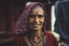 An elderly woman with a warm smile, wearing a patterned headscarf and a red cardigan. She has a nose ring and a bindi on her forehead, with visible wrinkles symbolizing age and experience. The background is blurred, suggesting an outdoor setting.