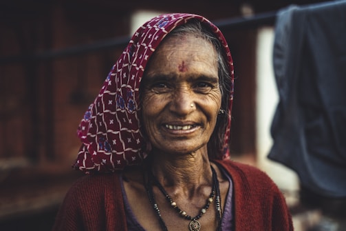 An elderly woman with a warm smile, wearing a patterned headscarf and a red cardigan. She has a nose ring and a bindi on her forehead, with visible wrinkles symbolizing age and experience. The background is blurred, suggesting an outdoor setting.
