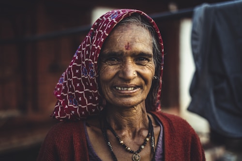 An elderly woman with a warm smile, wearing a patterned headscarf and a red cardigan. She has a nose ring and a bindi on her forehead, with visible wrinkles symbolizing age and experience. The background is blurred, suggesting an outdoor setting.