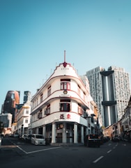 A heritage building with art deco architecture stands prominently at a street corner, featuring red accents and the number 1939 at the top. Modern skyscrapers with unique designs rise in the background, contrasting with the historical charm of the street. Automobiles line the narrow road leading toward the buildings.