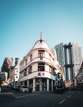 A heritage building with art deco architecture stands prominently at a street corner, featuring red accents and the number 1939 at the top. Modern skyscrapers with unique designs rise in the background, contrasting with the historical charm of the street. Automobiles line the narrow road leading toward the buildings.