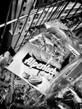 A close-up image of a shopping cart containing various products, including a jar of Alpenliebe Gold candies with its label prominently displayed. The scene is monochromatic and taken indoors, capturing a part of the metal grid of the cart and some packaging details.