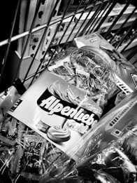 A close-up image of a shopping cart containing various products, including a jar of Alpenliebe Gold candies with its label prominently displayed. The scene is monochromatic and taken indoors, capturing a part of the metal grid of the cart and some packaging details.
