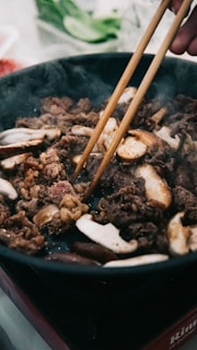 A pair of chopsticks is being used to cook a mixture of meat and mushrooms in a pan. The scene suggests a cooking process taking place with prominent steam rising from the dish. Vegetables are seen in the blurred background.