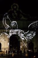 A large, illuminated projection of a winged figure holding a sword is displayed on the facade of a historical archway at night. The projection appears to depict an angel or a similar mythological character, with detailed wings and a dynamic pose. Below, people are gathered in the dimly lit street, and a car is parked to the side. The scene is atmospheric with a play of light and shadow on the architecture, adding a dramatic effect.