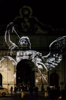 A large, illuminated projection of a winged figure holding a sword is displayed on the facade of a historical archway at night. The projection appears to depict an angel or a similar mythological character, with detailed wings and a dynamic pose. Below, people are gathered in the dimly lit street, and a car is parked to the side. The scene is atmospheric with a play of light and shadow on the architecture, adding a dramatic effect.