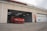 A red sports car is parked in front of an open garage bay at a racing circuit. The garage has a beige exterior with a large number 04 painted on the wall beside a closed garage door. Overhead, fluorescent lights illuminate the interior.