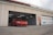 A red sports car is parked in front of an open garage bay at a racing circuit. The garage has a beige exterior with a large number 04 painted on the wall beside a closed garage door. Overhead, fluorescent lights illuminate the interior.