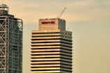 Two modern high-rise buildings with different architectural designs stand prominently. The tower on the right displays the word 'MAPFRE' on its top, with a crane visible on the roof, indicating construction or maintenance activities. The other building on the left features a unique metal frame structure on its facade.