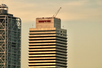 Two modern high-rise buildings with different architectural designs stand prominently. The tower on the right displays the word 'MAPFRE' on its top, with a crane visible on the roof, indicating construction or maintenance activities. The other building on the left features a unique metal frame structure on its facade.