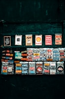 A collection of colorful vintage-style signs and magnets displayed on a dark green background. The signs feature various slogans, brand logos, and humorous phrases. They are arranged in rows, each with bright and contrasting colors.