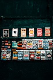 A collection of colorful vintage-style signs and magnets displayed on a dark green background. The signs feature various slogans, brand logos, and humorous phrases. They are arranged in rows, each with bright and contrasting colors.