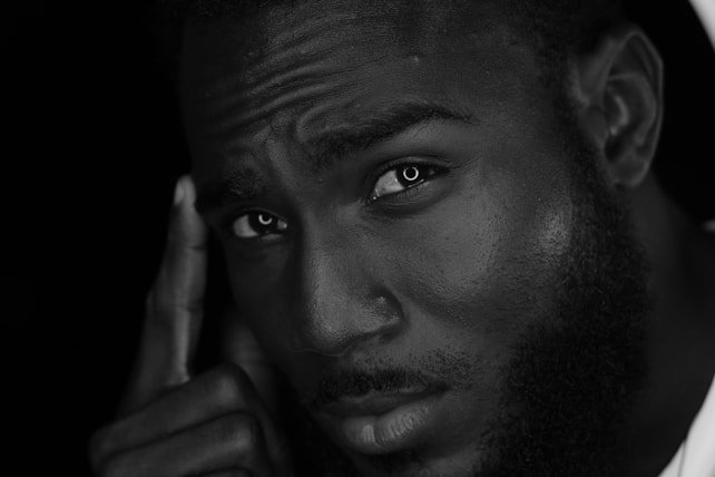 The black and white image depicts a close-up portrait of a man with a thoughtful expression. His eyes are piercing and his brow is furrowed as he holds his index finger to his temple. The lighting highlights the texture of his skin and beard.