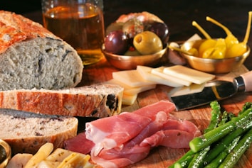 A rustic wooden table is laid out with various foods including slices of crusty bread, cured meat, steamed asparagus, different types of cheese, green and black olives, and bright yellow peppers. There is also a knife, a glass of what appears to be a light-colored liquid, possibly beer or cider in the background.
