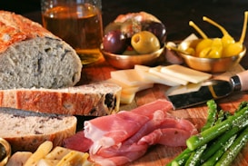 A rustic wooden table is laid out with various foods including slices of crusty bread, cured meat, steamed asparagus, different types of cheese, green and black olives, and bright yellow peppers. There is also a knife, a glass of what appears to be a light-colored liquid, possibly beer or cider in the background.