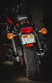 A motorcycle with a Honda logo visible on the seat is positioned in a dimly lit environment. The license plate has several labels, including one from San Francisco and a Massachusetts sticker. The vehicle is equipped with indicators, large rear suspension, and a prominent rear tire showcasing its tread pattern.