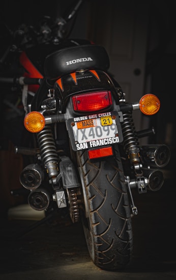 A motorcycle with a Honda logo visible on the seat is positioned in a dimly lit environment. The license plate has several labels, including one from San Francisco and a Massachusetts sticker. The vehicle is equipped with indicators, large rear suspension, and a prominent rear tire showcasing its tread pattern.