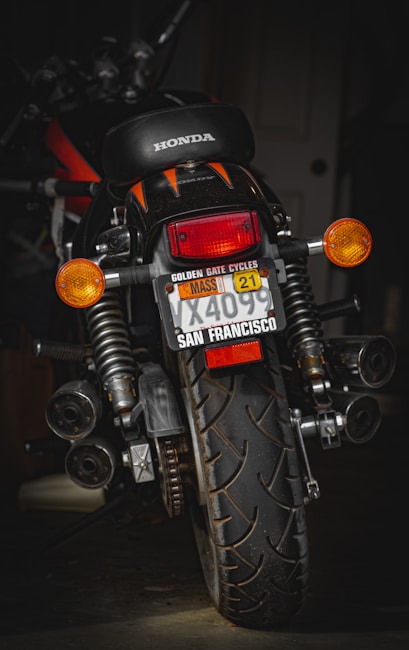 A motorcycle with a Honda logo visible on the seat is positioned in a dimly lit environment. The license plate has several labels, including one from San Francisco and a Massachusetts sticker. The vehicle is equipped with indicators, large rear suspension, and a prominent rear tire showcasing its tread pattern.