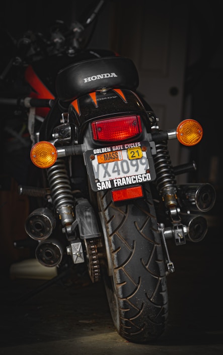 A motorcycle with a Honda logo visible on the seat is positioned in a dimly lit environment. The license plate has several labels, including one from San Francisco and a Massachusetts sticker. The vehicle is equipped with indicators, large rear suspension, and a prominent rear tire showcasing its tread pattern.