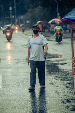 A person wearing a mask and cap walks on a wet street with puddles and reflections. In the background, there are motorcycles, some with riders wearing raincoats. Colorful umbrellas are visible along the roadside.