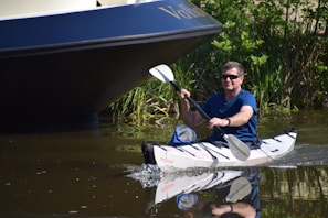 A person wearing sunglasses is kayaking on a calm body of water, with an overhanging part of a larger boat visible in the background and greenery along the shoreline.