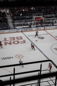 A hockey game is taking place in an indoor arena. Players wearing red uniforms are skating on the ice rink while a referee in a black and white striped uniform is present on the ice. There are a few spectators seated in the bleachers, and the boards are lined with advertisements. The ice features various markings and logos.