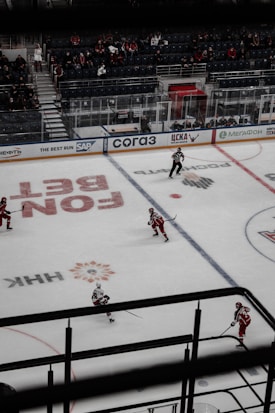 A hockey game is taking place in an indoor arena. Players wearing red uniforms are skating on the ice rink while a referee in a black and white striped uniform is present on the ice. There are a few spectators seated in the bleachers, and the boards are lined with advertisements. The ice features various markings and logos.