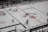A hockey game is taking place in an indoor rink. Players in red and white jerseys are actively engaged in play, skating across the ice towards the goal. The rink is marked with various logos and lines signifying boundaries and zones. Positioned around the rink are referees dressed in black and white stripes, and spectators can be seen behind the glass panels.