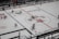 A hockey game is taking place in an indoor rink. Players in red and white jerseys are actively engaged in play, skating across the ice towards the goal. The rink is marked with various logos and lines signifying boundaries and zones. Positioned around the rink are referees dressed in black and white stripes, and spectators can be seen behind the glass panels.
