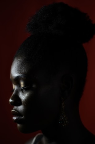 A profile view of a person with a dark complexion against a dark red background. The hair is styled in a puff with natural texture. The person has closed eyes with subtle makeup and wears a dangling earring.