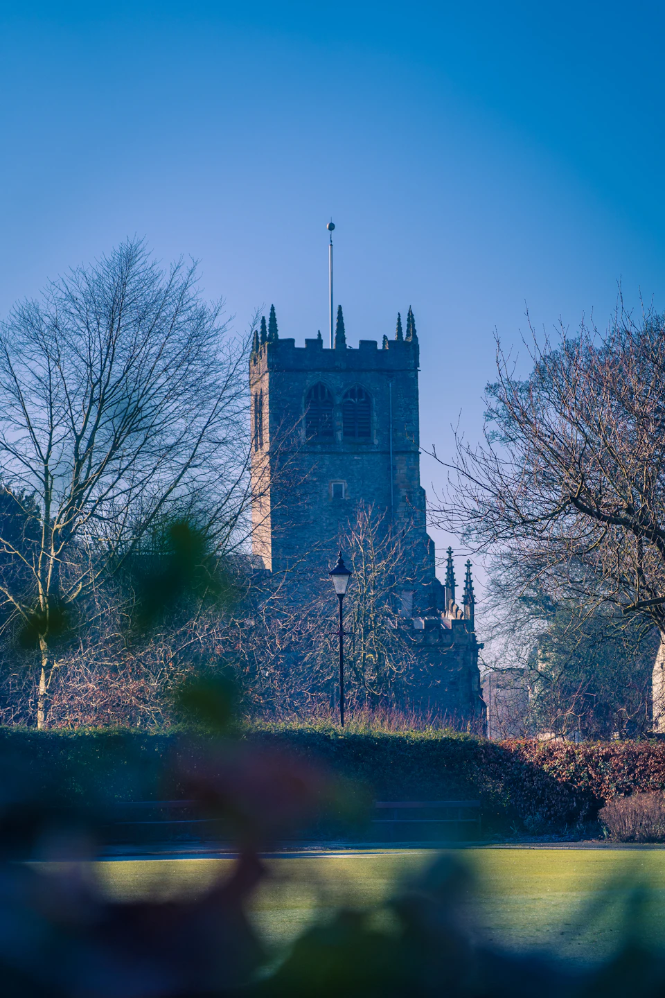 The image depicts a serene scene at a church, with its tall steeple reaching towards the clear blue sky. The church is nestled amidst nature, surrounded by trees and bushes that add to the peaceful atmosphere. In front of the church, there's a small pond reflecting the surrounding greenery. A solitary street lamp stands on the right side of the image, adding an element of human presence in this tranquil setting.