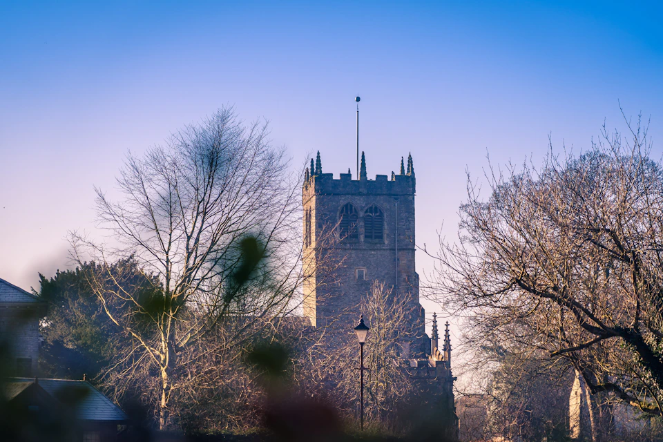 The image depicts a serene scene featuring a tall, stone church with a pointed roof and two towers standing against a clear blue sky. The church is surrounded by trees, adding to its peaceful ambiance. In front of the church, there are several bare-branched trees that contrast with the lush greenery surrounding them.

In the foreground, there's a street lamp positioned on the right side of the image, and a building can be seen in the background. The overall color palette is dominated by shades of green from the trees and browns from the stone church and buildings.