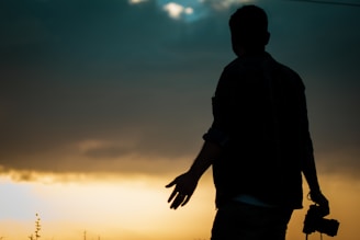 A silhouette of a person holding a camera against a dramatic sunset sky. The horizon glows with warm yellow and orange hues, contrasting with the darkened figure. The sky shows a gradient from light to dark blue as it reaches the top.