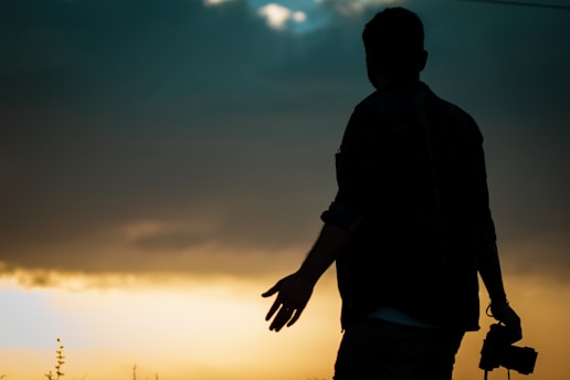 A silhouette of a person holding a camera against a dramatic sunset sky. The horizon glows with warm yellow and orange hues, contrasting with the darkened figure. The sky shows a gradient from light to dark blue as it reaches the top.