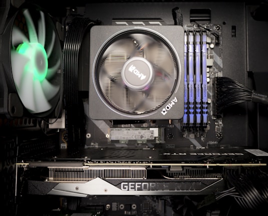 A computer interior showcasing key components such as a cooling fan with RGB lighting, a large central fan branded with 'AMD', multiple RAM sticks, and a visible graphics card labeled 'GEFORCE RTX'. Several cables are neatly arranged, and the motherboard is slightly visible.