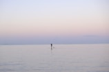 A lone individual engages in stand-up paddleboarding on a vast, calm, and peaceful body of water. The serene sky gradually transitions from soft pinks to pale blues, suggesting an early morning or late evening setting.