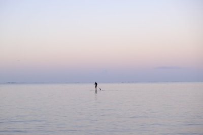 A lone individual engages in stand-up paddleboarding on a vast, calm, and peaceful body of water. The serene sky gradually transitions from soft pinks to pale blues, suggesting an early morning or late evening setting.