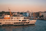 A ferry is cruising on a calm body of water during sunset. The skyline in the background features a cityscape with historic architecture, including minarets and domed buildings. The sky is tinted with warm hues, and a bustling waterfront filled with people and buildings appears on the right.