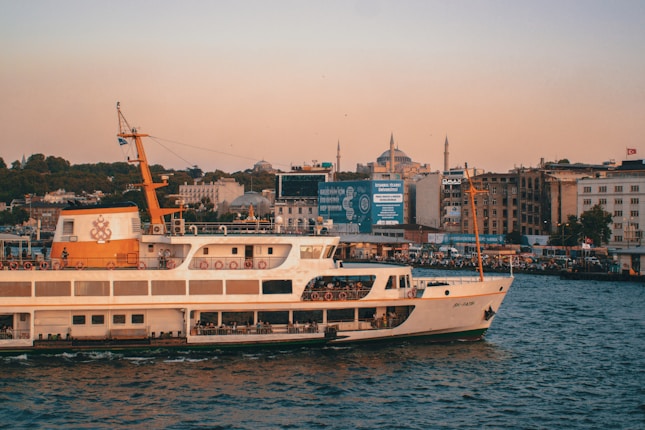 A ferry is cruising on a calm body of water during sunset. The skyline in the background features a cityscape with historic architecture, including minarets and domed buildings. The sky is tinted with warm hues, and a bustling waterfront filled with people and buildings appears on the right.