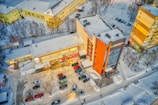 An aerial view of a parking lot adjacent to commercial buildings, with snow-covered streets and rooftops. Cars are parked in rows, and there's a clear path showing vehicular movement. Leafless trees surround the area, indicating winter season.