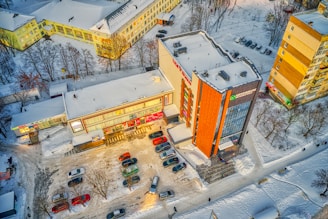 An aerial view of a parking lot adjacent to commercial buildings, with snow-covered streets and rooftops. Cars are parked in rows, and there's a clear path showing vehicular movement. Leafless trees surround the area, indicating winter season.