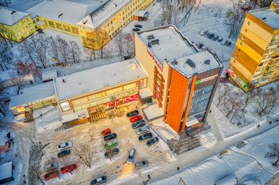 An aerial view of a parking lot adjacent to commercial buildings, with snow-covered streets and rooftops. Cars are parked in rows, and there's a clear path showing vehicular movement. Leafless trees surround the area, indicating winter season.