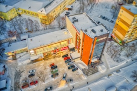 An aerial view of a parking lot adjacent to commercial buildings, with snow-covered streets and rooftops. Cars are parked in rows, and there's a clear path showing vehicular movement. Leafless trees surround the area, indicating winter season.